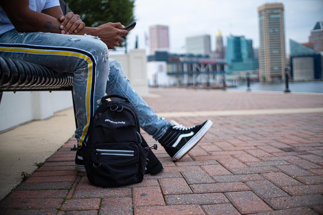 Person sitting in Baltimore's inner harbor, showing just their shoes and backpack on the pavement with the harbor in the background